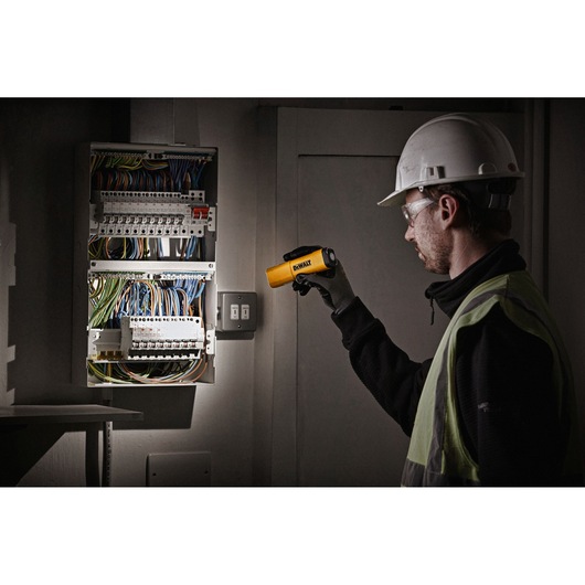A person wearing safety gear uses a DEWALT DCL183 portable work light to illuminate an electrical panel with multiple wires and switches.