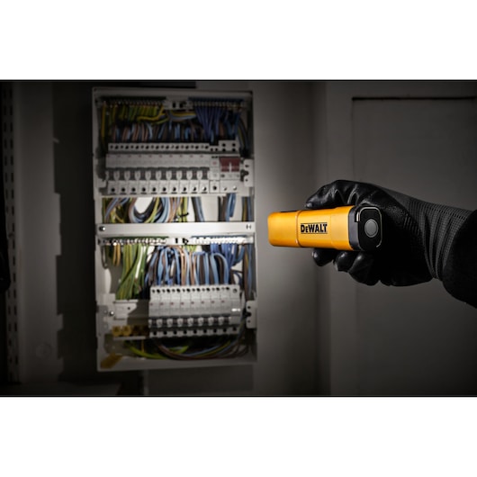 A person wearing a black glove is holding a yellow DEWALT flashlight, pointing it at an open electrical panel with multiple colored wires and circuit breakers.