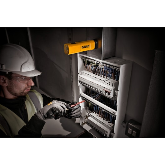 A DEWALT DCL183 portable LED work light is mounted above an open electrical panel, illuminating the workspace while a person in safety gear works with a screwdriver on the panel.