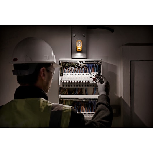 A person wearing a safety helmet and high-visibility vest is inspecting an electrical panel under the light of a mounted DEWALT DCL183 work lamp.