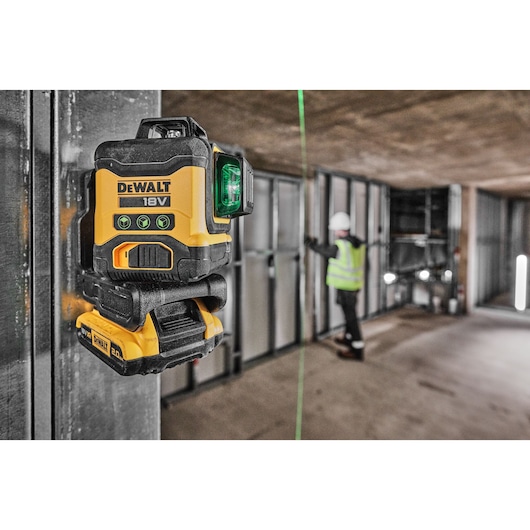 A DEWALT 18V green laser level mounted on a metal frame at a construction site. The laser projects a green line across the room, and a person wearing safety equipment is working in the background.