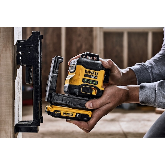 A person is holding a DEWALT 18V laser level with a 2 Ah battery, positioning it next to a wall-mounted bracket in an indoor construction setting.