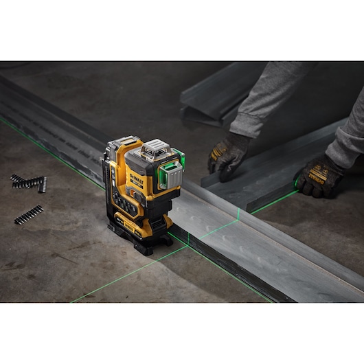 A DEWALT laser level tool projecting green laser lines on a construction site floor, with a person wearing gloves aligning metal pieces.