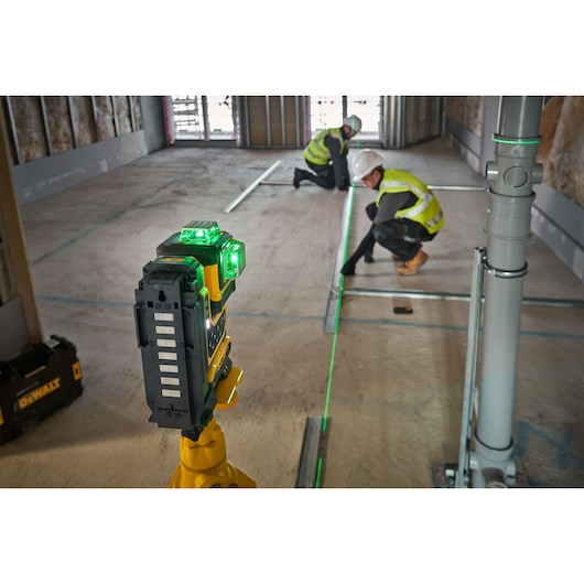 A DEWALT green laser level on a tripod projects a bright green line across a construction site floor, where two workers wearing high-visibility vests and helmets are aligning metal tracks and working on layout measurements.
