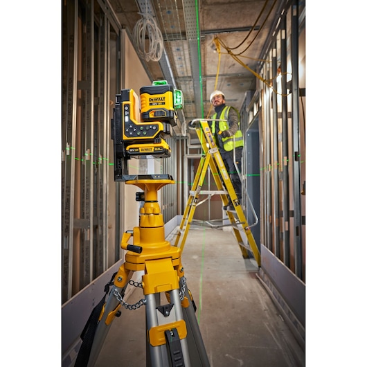 A DEWALT construction laser level (model DCLE34035D1) is mounted on a tripod inside an unfinished building. The laser projects green lines across the walls and ceiling. In the background, a person wearing a safety vest and hard hat is working on a ladder.