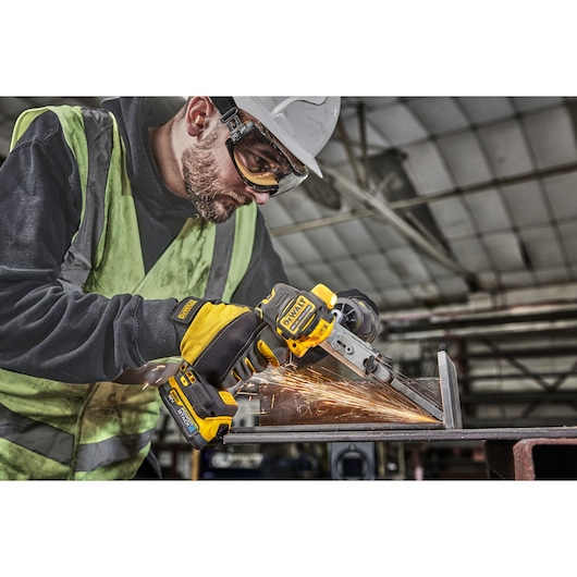 A worker wearing protective gloves, a safety vest, and a helmet is using a DEWALT cordless power tool to grind or cut a metal piece, with sparks flying in an industrial setting.