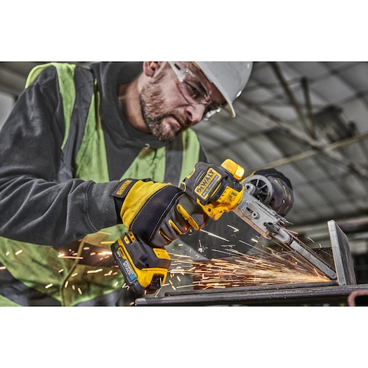 A worker wearing gloves and a safety vest uses a DEWALT cordless power tool to grind or sand a piece of metal, creating sparks in an industrial setting.