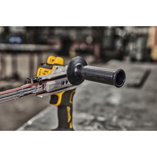 Close-up of a DEWALT cordless electric belt file tool, featuring a side handle and abrasive belt. The tool is set on a workbench in an industrial workshop setting.
