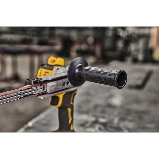 Close-up view of a DEWALT cordless belt sander (model DCM200NT) with a black auxiliary handle, placed on a workbench in an industrial setting.