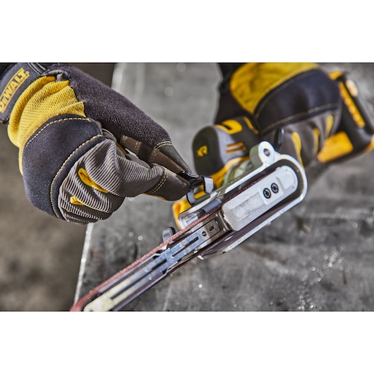Close-up of a person wearing DEWALT work gloves adjusting a DEWALT cordless power tool, possibly a belt sander, on a workbench.
