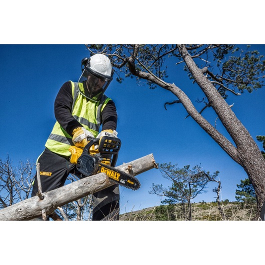 A person wearing safety gear is using a DEWALT chainsaw to cut a tree branch outdoors. The sky is clear and trees are visible in the background.