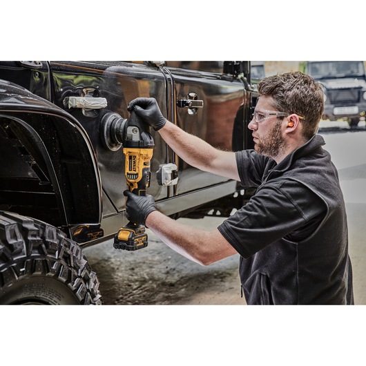 A person wearing black gloves uses a DEWALT cordless polisher (model DCM848N) to polish the side of a black off-road vehicle. The scene takes place in a workshop or garage.