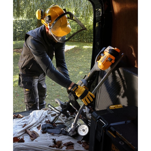 A person wearing safety gear, including a yellow helmet and gloves, is handling a DEWALT cordless outdoor power tool inside a vehicle. The tool is resting among other equipment and leaves on a cloth, with a green outdoor area visible in the background.