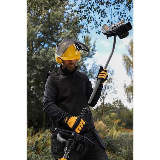 Person wearing safety gear and gloves operates a DEWALT outdoor tool in a grassy area with trees and sky in the background.