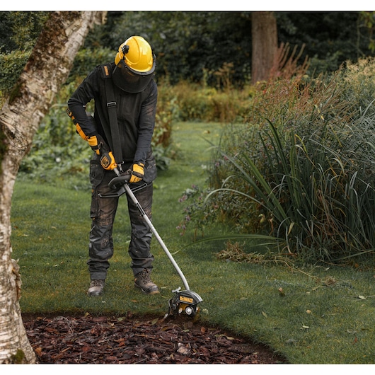 A person wearing protective gear uses a DEWALT lawn edger tool to trim the edge of a garden bed in a grassy outdoor area.
