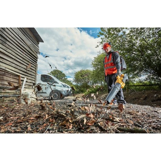 A person wearing safety gear and a red vest uses a DEWALT leaf blower outdoors to clear leaves and debris from a gravel area. There is a white van, a wooden building, and trees in the background.