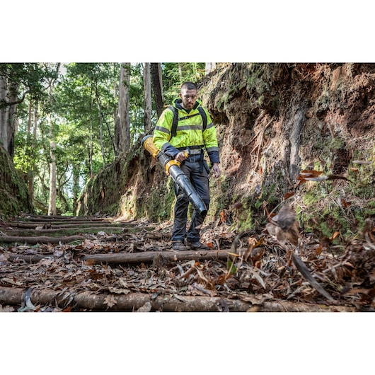 A person wearing a high-visibility jacket is using a DEWALT leaf blower to clear fallen leaves from a wooded path. The face of the person is blurred for privacy.