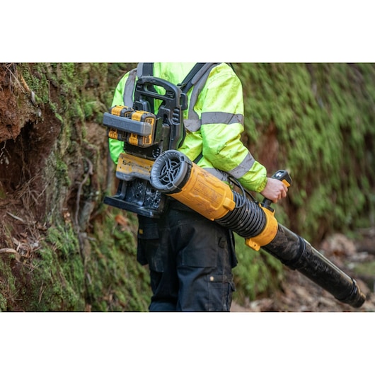 A person wearing a high-visibility jacket is using a DEWALT cordless backpack blower outdoors, walking along a mossy, wooded area.