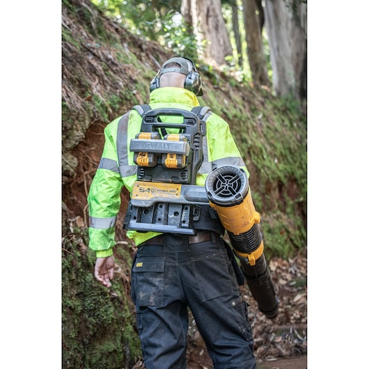 A person wearing a high-visibility jacket and ear protection operates a DEWALT backpack leaf blower in an outdoor wooded area. The DEWALT device is visible, with batteries and blower tube attached.