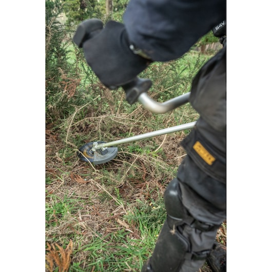 A person using a DEWALT brush cutter (model DCMBC723N) to trim dense vegetation outdoors. The user is wearing protective gloves and clothing while operating the power tool in a grassy area.