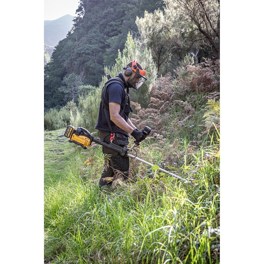 A person wearing protective gear and gloves uses a DEWALT DCMBC823N battery-powered brush cutter to trim dense grass and plants outdoors in a natural, hilly landscape.