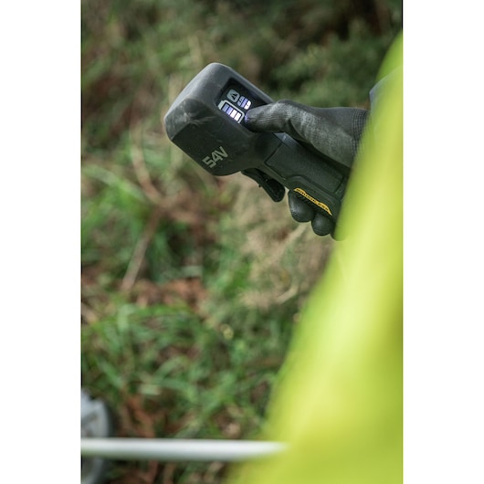 Close-up of a gloved hand holding a DEWALT 54V cordless tool with a digital display. The background shows outdoor greenery.