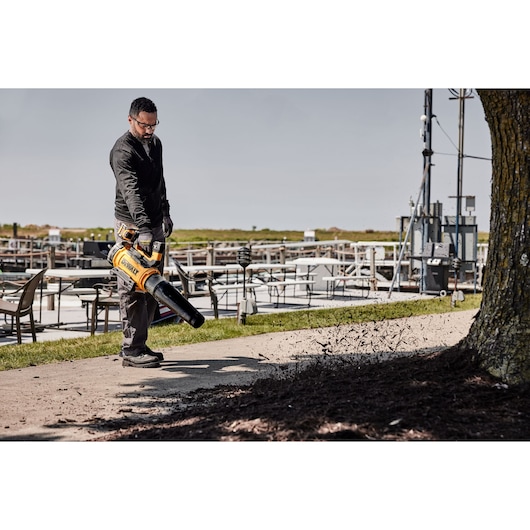 A person uses a DEWALT cordless leaf blower (model DCMBL777N) to clear leaves and debris from an outdoor pathway near a tree, with outdoor seating and equipment visible in the background.