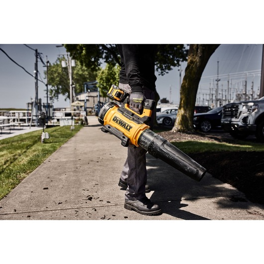A person using a DEWALT cordless leaf blower on a sidewalk, clearing debris. The leaf blower is prominently yellow and black with visible DEWALT branding.