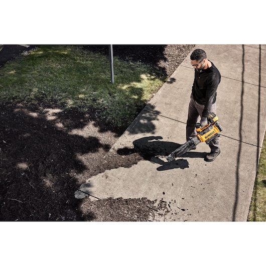 A person using a DEWALT cordless leaf blower (model DCMBL777N) to clear debris from a concrete sidewalk next to a grassy area and mulch.