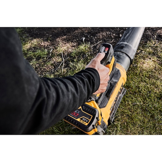 A person using a DEWALT cordless leaf blower with an 18V FlexVolt battery, outdoors on grass near a patch of dirt.