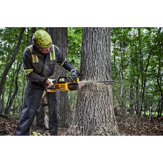 A person wearing protective gear is using a DEWALT cordless chainsaw (model DCMCS575N) to cut down a large tree in a forest.