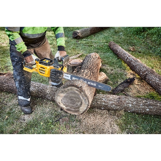 A person using a DEWALT cordless chainsaw to cut a log outdoors, with wood chips flying and several tree trunks on the grass.
