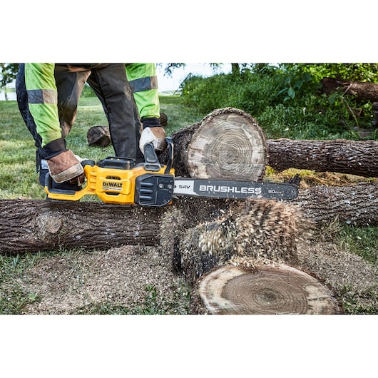 A person using a yellow DEWALT cordless brushless chainsaw to cut a thick log outdoors. Sawdust is flying as the chainsaw cuts through the wood.