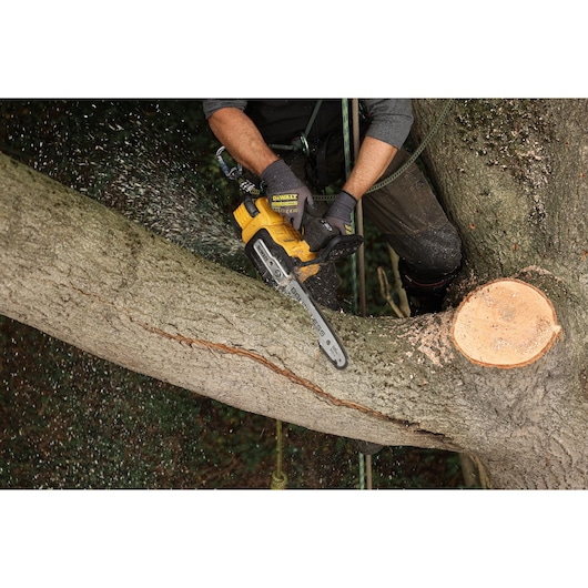 A person using a DEWALT cordless chainsaw to cut a tree branch outdoors. Sawdust is flying as the branch is being trimmed.