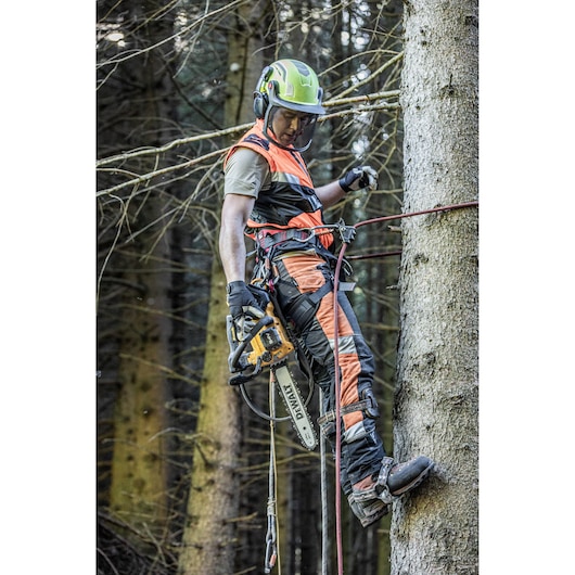 A person wearing safety gear and climbing equipment operates a DEWALT chainsaw while secured to a tree in a forest setting. The individual is equipped with a helmet, gloves, and protective clothing. The DEWALT chainsaw is prominently visible.