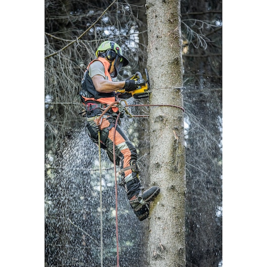 A person wearing protective gear uses a DEWALT chainsaw to cut a tree while suspended with safety ropes. Sawdust is flying as the chainsaw operates.