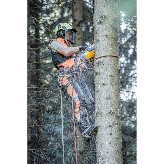A person wearing safety gear and climbing equipment uses a yellow DEWALT chainsaw to cut a tree trunk in a forest, with wood chips flying during operation.