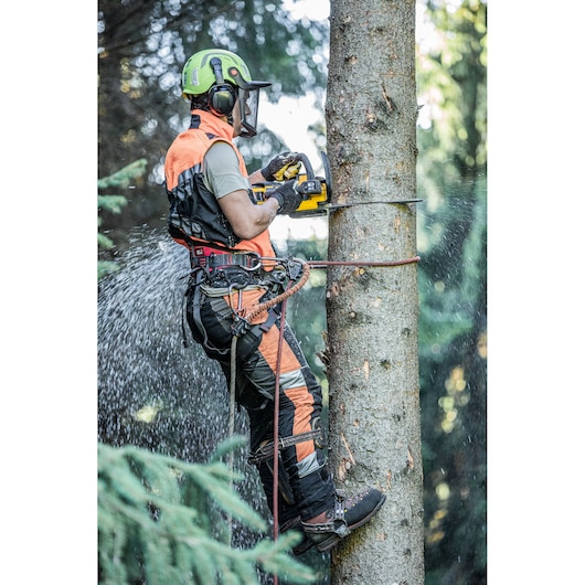 A person wearing safety gear, including a helmet, ear protection, and harness, uses a DEWALT chainsaw to cut a tree trunk while secured with ropes. Wood chips are flying as the saw cuts into the tree.