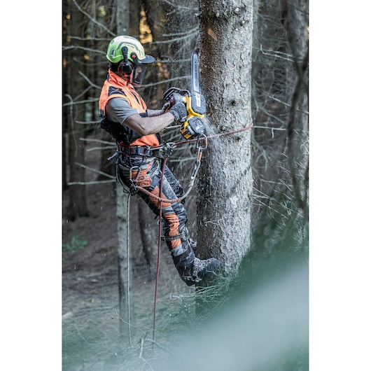 A person wearing safety gear uses a DEWALT chainsaw to cut a tree while suspended by ropes in a forest.