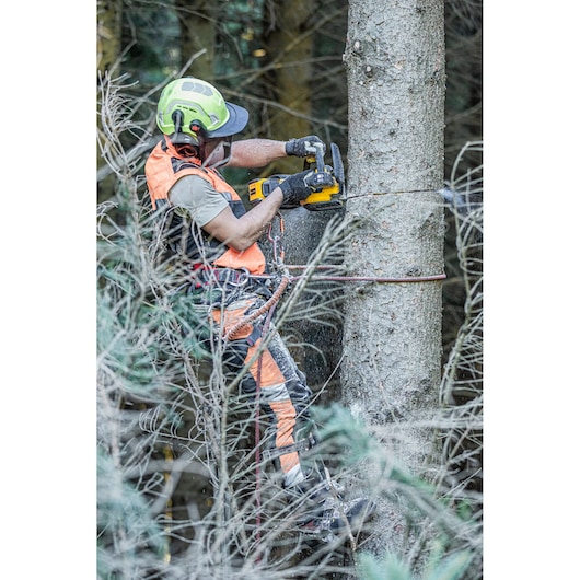 A person wearing safety gear uses a DEWALT cordless chainsaw to cut a tree in a forest. They are secured with climbing equipment and are surrounded by branches.