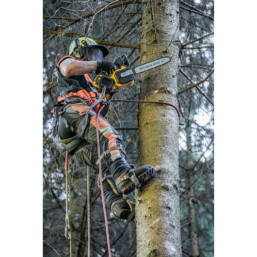 A person wearing safety gear is using a DEWALT cordless brushless chainsaw to cut branches while climbing a tree in a forest. The individual is equipped with harnesses, a helmet, and protective clothing.