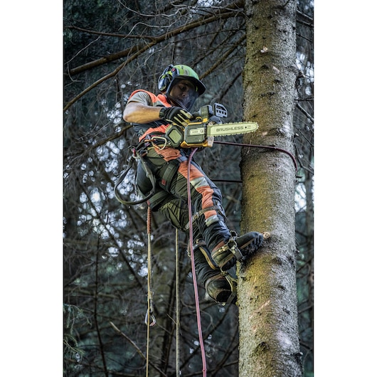 A worker in protective gear is climbing and cutting a tree using a DEWALT brushless chainsaw. He is wearing safety equipment and is secured with ropes in a forest setting.