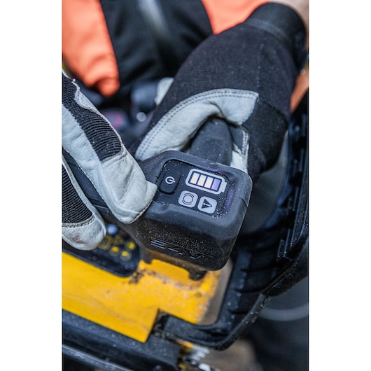 Close-up of a gloved hand operating the control panel of a DEWALT cordless chainsaw, showing battery level indicators and control buttons.