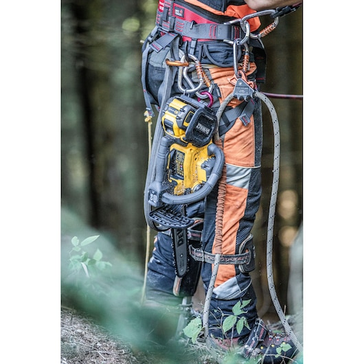 A close-up of a person wearing safety gear and a harness in a forest, with a DEWALT cordless chainsaw attached to their belt.
