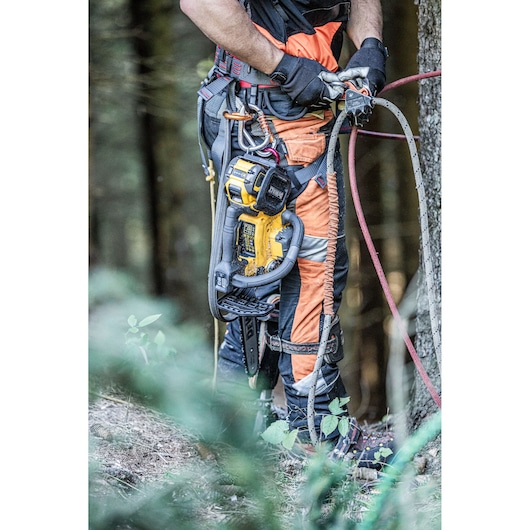 A person wearing protective orange and black work clothing is using climbing gear and holding ropes next to a tree in a forest. A DEWALT cordless chainsaw is secured to the person's harness.