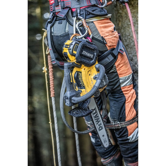 A close-up of a DEWALT cordless chainsaw (model DCMCST635X1) attached to the harness of a person wearing safety gear and climbing equipment, with trees in the background.