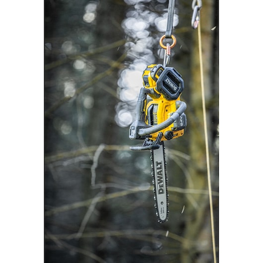 A DEWALT cordless chainsaw is hanging from a rope and hook in an outdoor forest setting. The saw is yellow with black detailing and features a visible DEWALT logo on the blade.