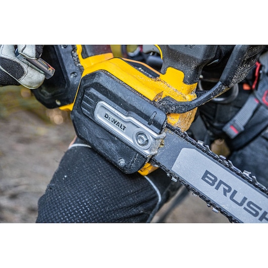 Close-up view of a DEWALT chainsaw in use, showing the detail of the chainsaw chain and the brand label on the body. The chainsaw is being operated outdoors by a person wearing gloves and protective clothing.