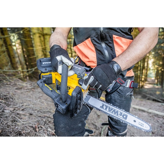 A person wearing protective gloves and gear is holding a DEWALT cordless chainsaw in a forest setting.