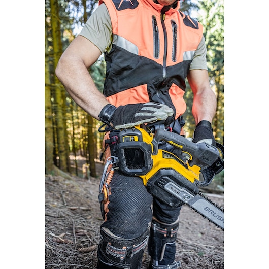 A person wearing protective clothing and gloves is operating a DEWALT cordless chainsaw in a forest setting.